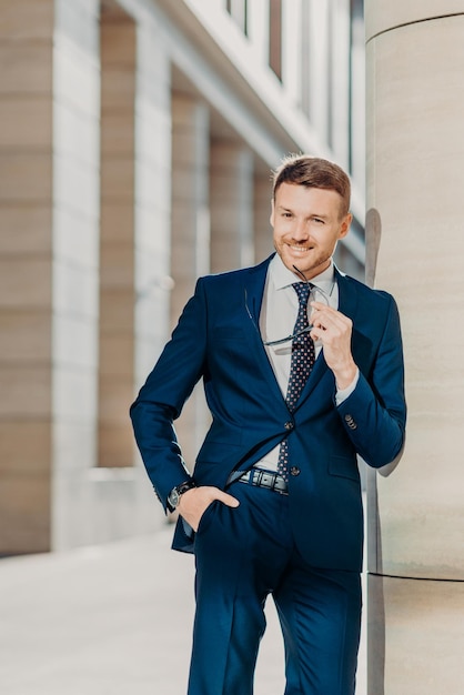 Thoughtful cheerful bearded male entrepreneur has pleasant
smile wears black suit holds sunglasses poses indoor being
delighted with successful business meeting and signing contract
with partner