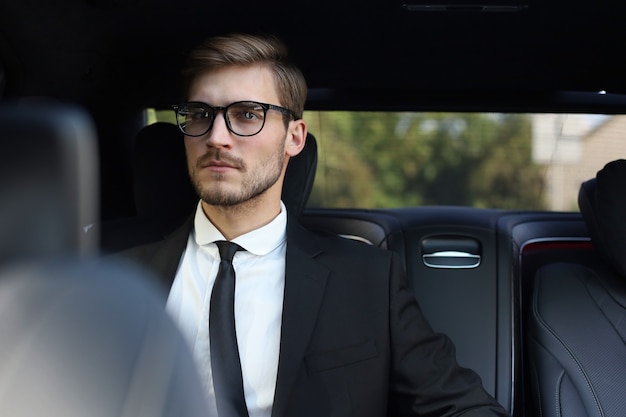 Thoughtful confident businessman in full suit with eyewear looking away while sitting in the car.