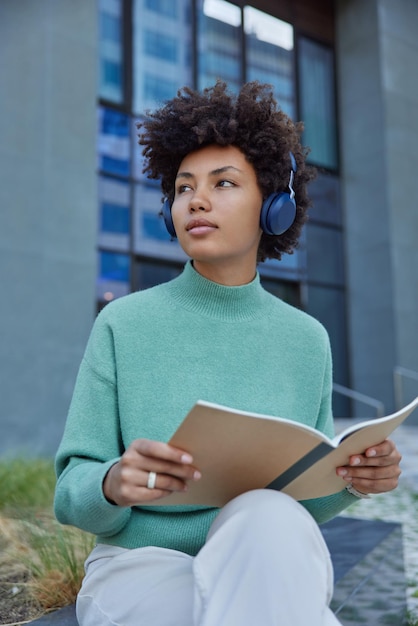 Thoughtful curly female student listens educational webinar in wireless headphones concentrated away pensively holds opened notepad wears casual jumper and trousers poses outside in urban place