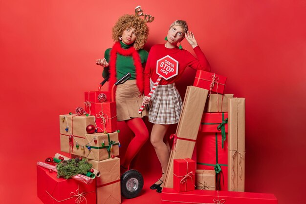 Thoughtful displeased women being busy decorating house before\
new year wear festive clothes pose around stacks of wrapped\
presents isolated over red background friendship and holidays\
concept