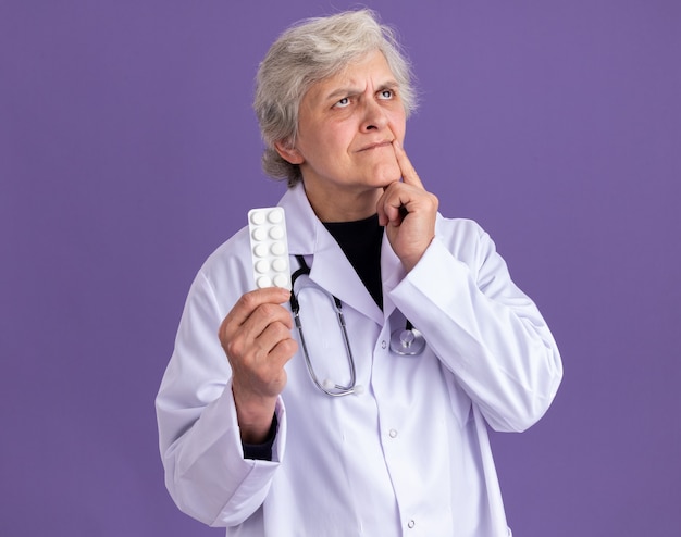 thoughtful elderly woman in doctor uniform with stethoscope holding pill packaging and looking up isolated on purple wall