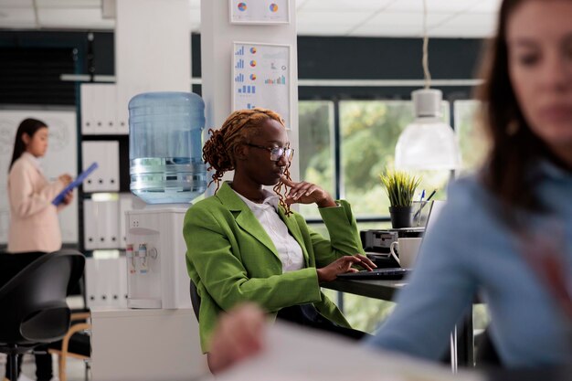 Thoughtful employee working business office open space, manager sitting at workplace desk, colleagues on background. Young woman thinking, typing on computer, side view medium shot