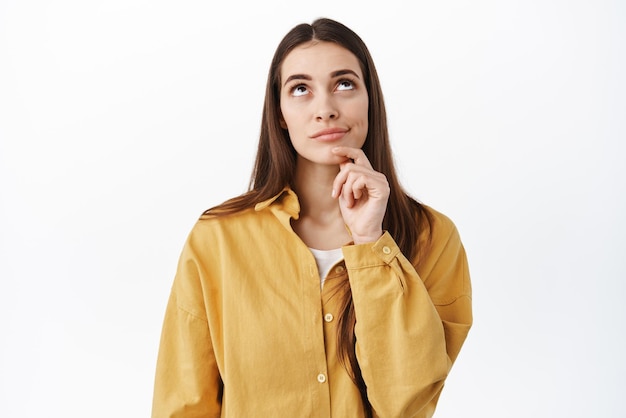 Thoughtful female model touch chin looking up at top promo deal and thinking making up her mind choosing standing pensive against white background