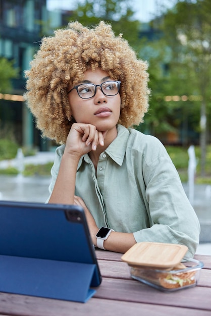 Thoughtful female student watches training webinar via touch\
pad sits at wooden table outdoors eats lunch wears spectacles linen\
shirt and smartwatch reads information in internet technology\
concept