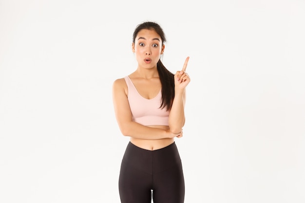 Thoughtful fit and slim asian girl have idea, sportswoman raise index finger for suggestion, have workout plan, standing white background.