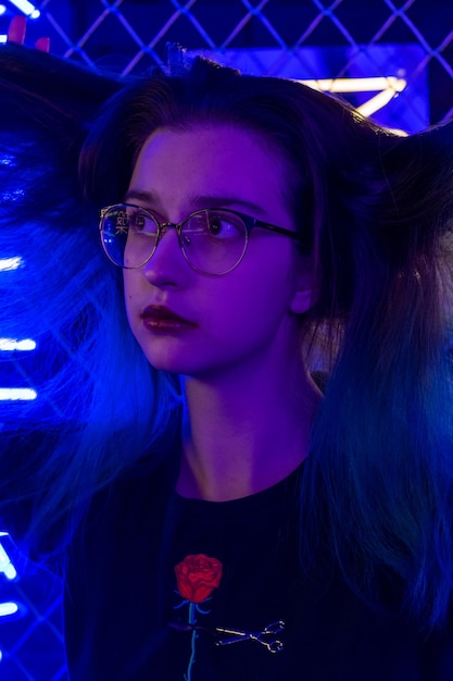 Thoughtful girl in glasses standing on the street near the evening neon lights Stylish trendy portrait of a girl in the style of the 80s