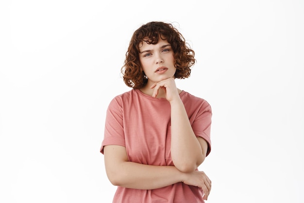 Thoughtful girl looking at camera pensive, touching chin and squinting suspicious, pondering something, standing against white background