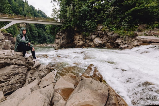 Thoughtful guy traveler sitting by a mountain stream in a\
cloudy forest and mountains