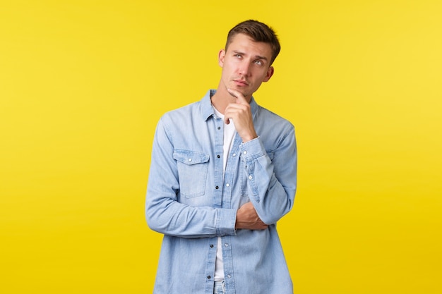 Thoughtful handsome adult blond guy thinking, calculating something in mind, rub chin and looking upper left corner doubtful, making choice in store, standing yellow background.
