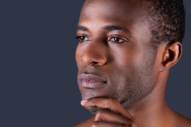 Thoughtful handsome. Young African man holding hand on chin while standing against black background