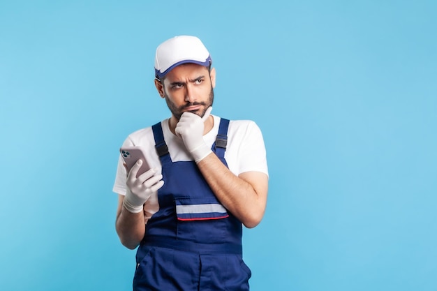 Thoughtful handyman in overalls and gloves holding mobile phone and pondering intensely, thinking over accepted online order as delivery, repair and maintenance services. indoor studio shot isolated