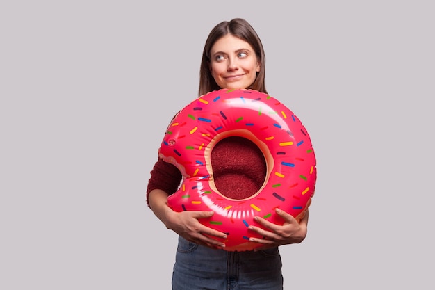 Thoughtful happy girl holding rubber ring pink donut and looking away with dreamy expression