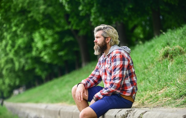 Вдумчивый хипстер сидит в парке, наслаждается природой этой весной, мужской модный стиль, он носит случайный брутальный хипстер с длинной бородой и крашеными волосами, зрелый студент, отдыхает на свежем воздухе, отдыхает
