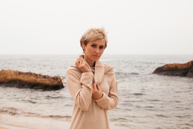 Thoughtful hipster woman with short haircut wearing dress near sea in autumn