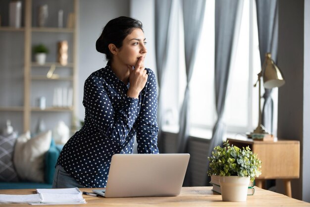 thoughtful indian woman standing at desk businesswoman freelancer pondering difficult task looking out window touching chin pensive young female student working on research project