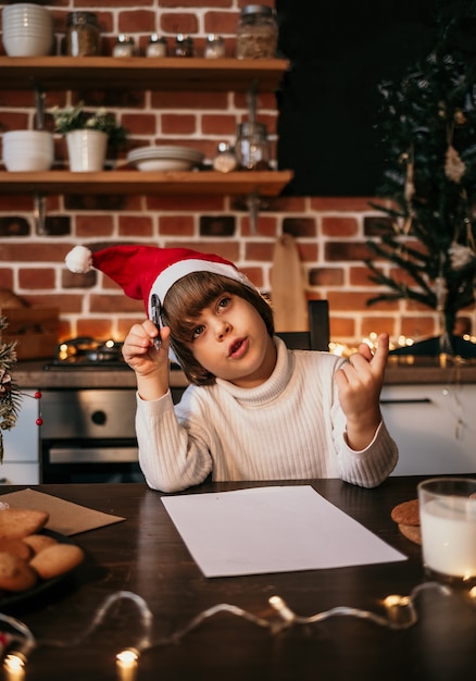 A thoughtful little boy in a white knitted sweater and a red hat writes a New Year's letter