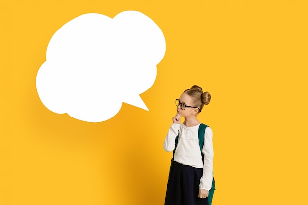 Photo thoughtful little schoolgirl looking at blank speech bubble and touching chin