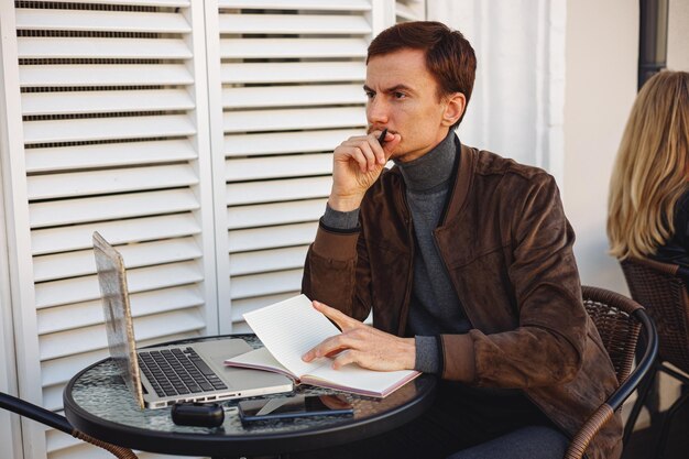 Thoughtful male freelancer with notebook and laptop leaning on hand and thinking in outdoor cafe