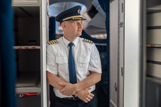 飛行機の中に立っている間、腕を組んで脇を向いたままの制服と帽子をかぶった思いやりのある男性パイロット。輸送、航空機搭乗員のコンセプト