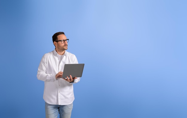 Foto professionale maschio riflessivo con computer wireless che guarda lontano e in piedi sullo sfondo blu