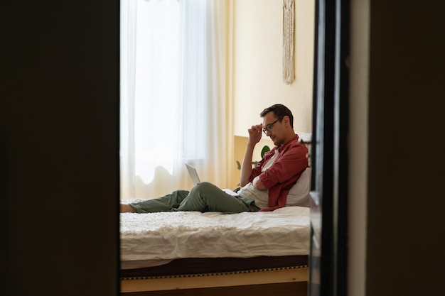 Thoughtful man freelancer sitting on bed with laptop stuck with task while working remotely