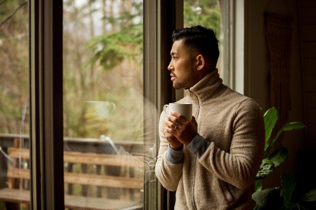 Photo thoughtful man holding coffee cup and looking through window at home