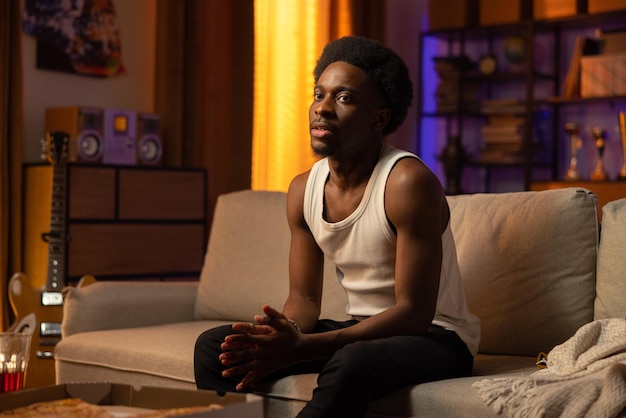 A thoughtful man sits on the sofa in the living room of a cozy apartment lit by the setting sun afro