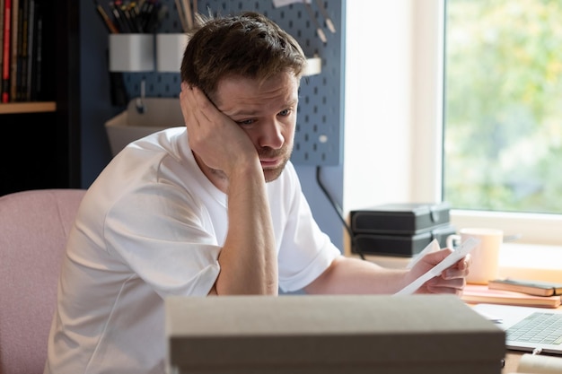 Thoughtful man sitting at table at home with papers trying to count his money and debts. Pensive confused young man thinking of problem solution