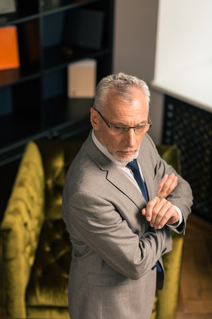 Thoughtful man standing with his hands crossed in his office near an armchair