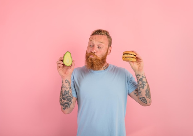 Thoughtful man with beard and tattoos is undecided if to eat an avocado or an hamburger