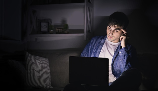 Thoughtful man with laptop at night. Young guy working late at home in light of computer screen