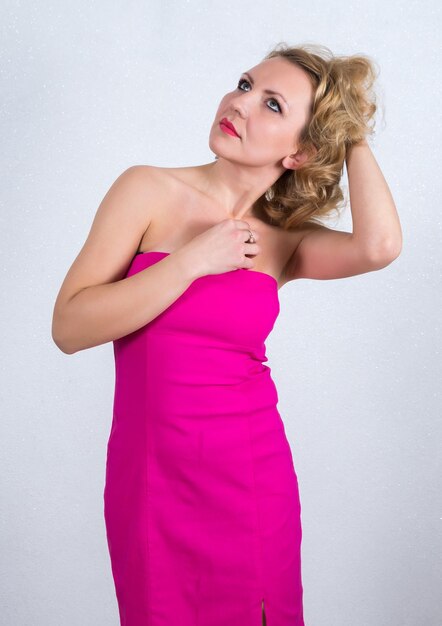 Thoughtful mid adult woman in pink off shoulder dress standing with hand in hair against gray background