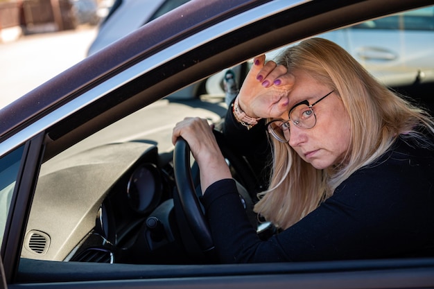 Thoughtful middle-aged blonde in glasses sits behind the wheel of a car view through the window