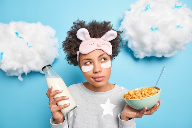 Thoughtful millennial girl has healthy breakfast holds bowl of cornflakes and milk bottle dressed in nightwear applies patches to reduce puffiness isolated over blue wall