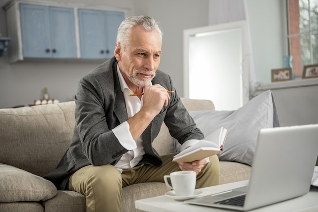 Thoughtful mood. Handsome male person keeping smile on his face and leaning elbows on knees while looking straight at screen of his laptop