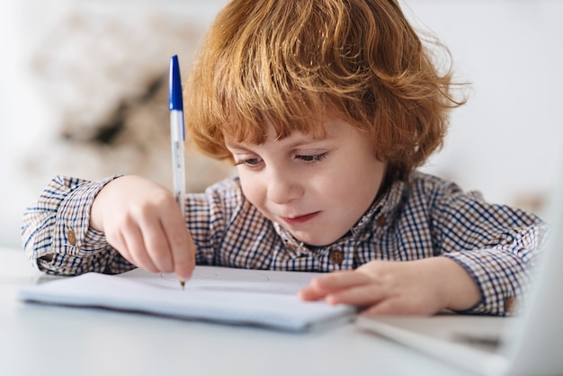 Mattina premurosa. affascinante ragazzo dai capelli rossi motivato che studia in una stanza illuminata dal sole mentre prende appunti e si siede al tavolo bianco