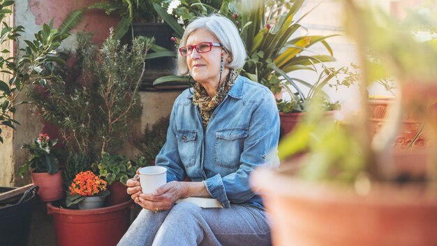 Thoughtful older woman sitting between plants holding cup