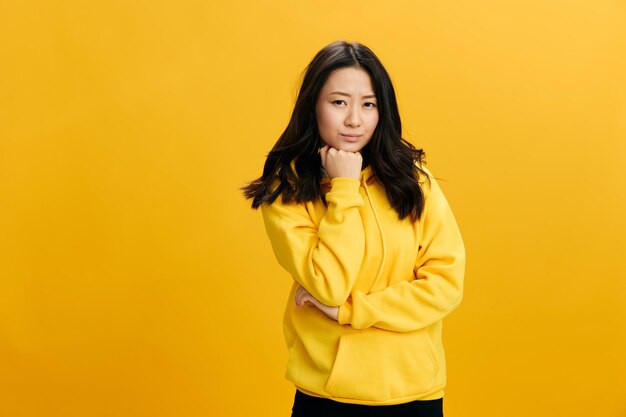 Thoughtful pensive lovely asian young woman in yellow hoodie\
sweatshirt can39t make difficult decision reclines on hand posing\
isolated on orange studio background cool offer people emotions\
concept