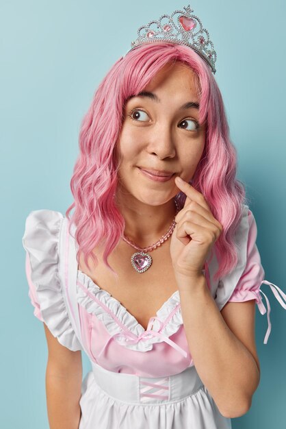 Thoughtful pink haired asian housewife keeps index finger near
corner of lips looks aside being deep in thoughts wears crown white
dress isolated on blue background thinks how to bring house in
order