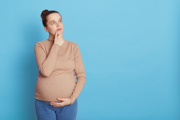 Thoughtful pregnant woman looks thoughtfully aside with hand on chin, makes planning about child birth