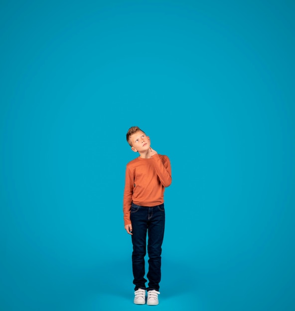 Photo thoughtful preteen boy looking up while standing over blue studio background