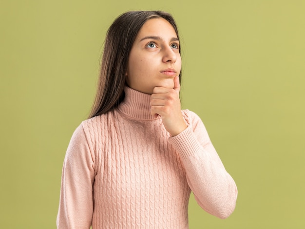 Thoughtful pretty teenage girl looking at side keeping hand on chin isolated on olive green wall with copy space