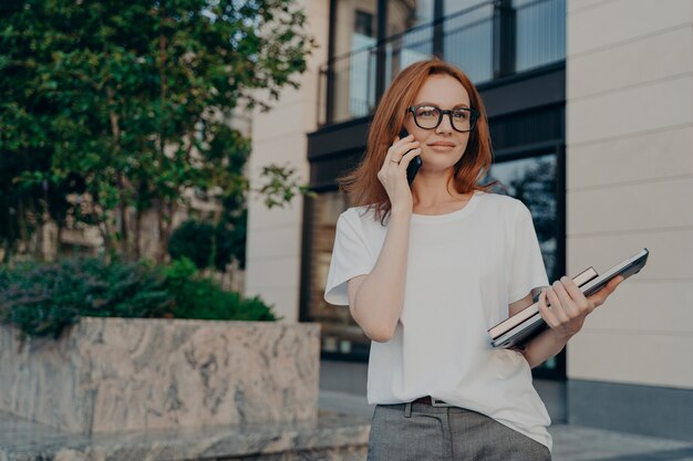 思いやりのある赤毛の女性が遠くを見ると、電話がスマートフォンを耳の近くに保持します