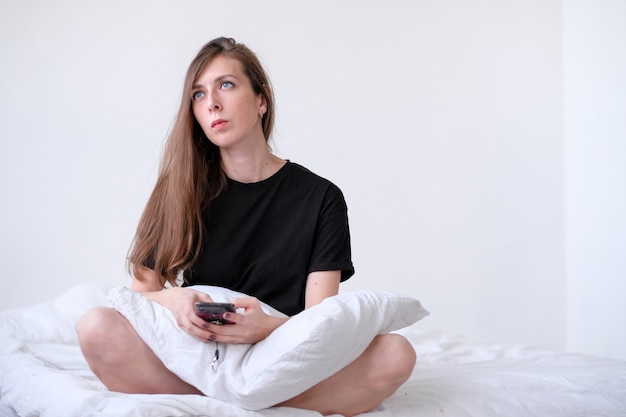 Thoughtful redhead young woman holding mobile phone and thinking in bed. young girl with blue eyes. sick leave with a phone. work at home