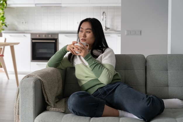 Задумчивая расслабленная азиатка с чашкой чая и кофе, мечтательно отводя взгляд, сидит на диване в гостиной
