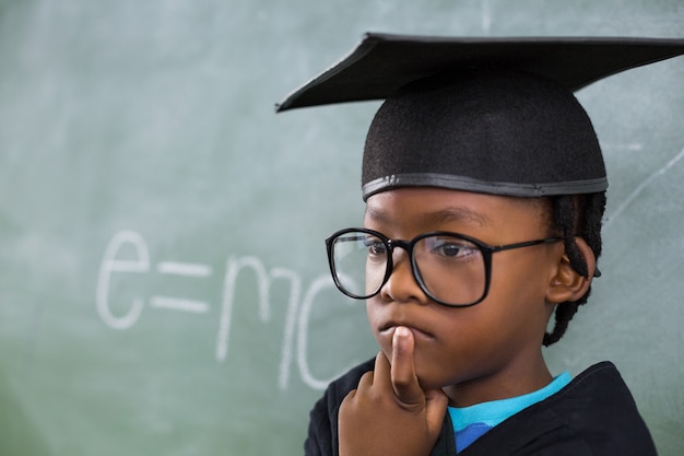 Thoughtful schoolboy wearing mortar board in classroom