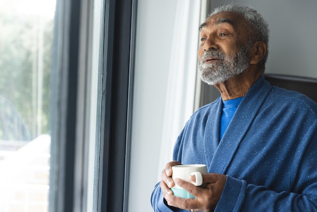 Thoughtful senior biracial man wearing bathrobe and holding mug of coffee by window at home