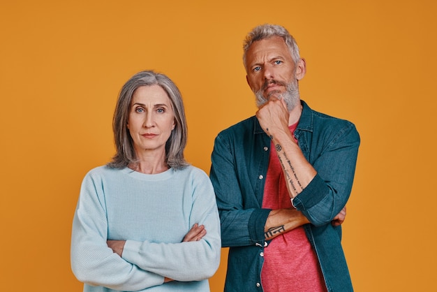 вдумчивый старший пара смотрит в камеру, стоя вместе на оранжевом фоне