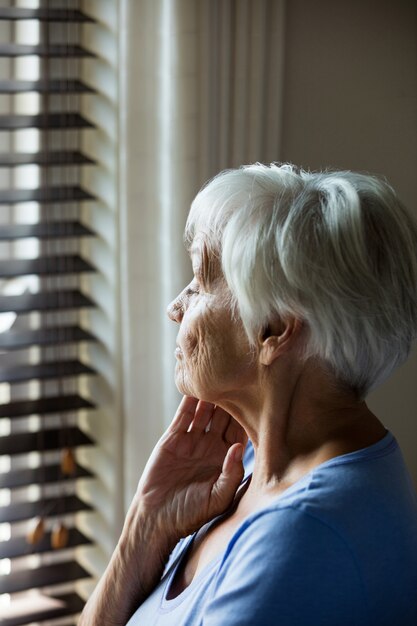 Задумчивая старшая женщина, выглядывающая из окна дома