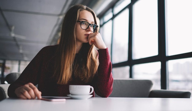 Thoughtful and serious girl looking in window while waiting colleague and having tea